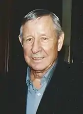 Portrait d'un homme âgé aux cheveux gris portant une veste noire sur une chemise bleue.