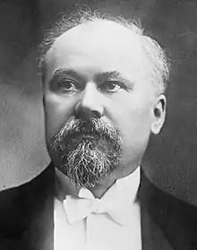 Photo en noir et blanc d'un homme au front dégarni, bouc et moustache poivre et sel, nœud papillon blanc sur col montant.