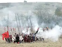 Photographie d'un groupe d'hommes au milieu d'un pré. Certains brandissent de longues piques, d'autres viennent de tirer des coups de feu qui ont généré un gros nuage de fumée blanche