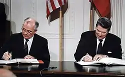 La photographie couleur montre les deux chefs d'état attablés pour une signature conjointe. Derrière eux, les drapeaux américain et soviétique ornent le mur.