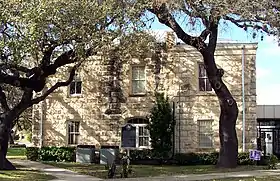 Vue d'un petit bâtiment à la façade de pierres blanches avec des arbres au premier plan.