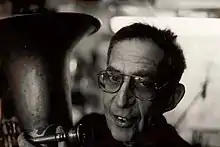 Photographie en noir et blanc d'un homme âgé. Il porte des lunettes et tient un tuba.