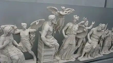 Une dizaine de statues blanches alignées : au centre un homme barbu, face à lui une femme en armes ; entre eux une figure féminine en vol ; divers autres personnages