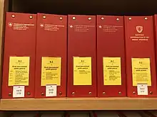 Photographie de cinq classeurs rouges avec étiquette jaune contenant le Recueil systématique sur une étagère en bois