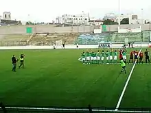 Dans un stade de football, des photographes vont sur le terrain pour photographier une équipe au maillot vert.