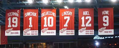 Photo couleur des 6 bannières rouges des maillots retirés des Red Wings de Détroit. De gauche à droite : Yzerman 19, Sawchuk 1, Delvecchio 10, Lindsay 7, Abel 12, Howe 9.