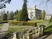 Un escalier de pierre menant à un petit jardin à la française en contrebas sur la gauche. Au loin sur la droite, derrière quelques arbres, se trouve un grand bâtiment cubique en pierres blanches, ressemblant à un manoir, avec de nombreuses cheminées et fenêtres