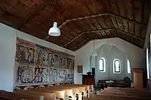 Photographie en couleurs d'une salle allongée, avec à gauche des scènes religieuses peintes sur fond blanc.