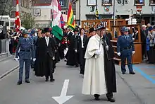 Photographie d'un groupe de personnes en costume sombre, encadrés par deux gendarmes en uniforme bleu ; devant, un homme en costume noir et blanc portant un sceptre ; derrière eux, plusieurs drapeaux portés.