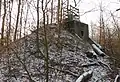 Photographie récente en couleurs d'une colline artificielle conique portant à son sommet une infrastructure bétonnée. L'ensemble est envahi par la forêt.
