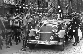Photo noir et blanc prise en septembre 1935. Dans une rue de Nuremberg, Adolf Hitler, debout à l’arrière d’une voiture au premier plan, salue des troupes de miliciens de la SA défilant sur sa gauche. On distingue aussi, à l’arrière de la voiture, juste derrière Hitler, le drapeau à croix gammée de cérémonie, Blutfahne, et, en uniforme SS, debout à droite, devant la portière de la voiture, Jakob Grimminger, porteur officiel du Blutfahne.