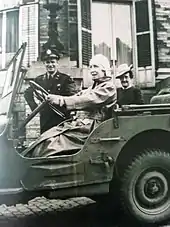 Assise au volant d'une Jeep, la reine vêtue d'un manteau en cuir et coiffée d'un casque se trouve devant un officier souriant et portant un képi et d'une dame coiffée d'un chapeau-cloche