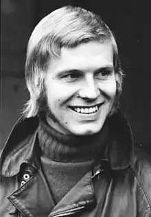 Photographie en noir et blanc, d'un homme de peau blanche, blond, vu de trois-quarts, souriant, en gros plan.