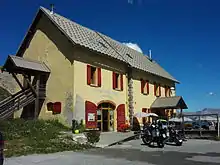 Façade et pignon d’une maison à un étage, murs jaunes et volets rouges.