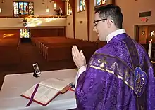 Un homme portant des vêtements violets et debout devant un autel utilise la caméra d'un téléphone portable pour s'enregistrer. Des bancs vides sont visibles à l'arrière-plan.