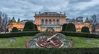 Des fleurs dessinent une horloge que des aiguilles parcourent. Derrière un grand bâtiment style demeure XIXème.