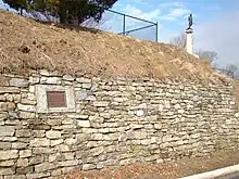 Photographie d'un mur de pierre avec un talus de terre au-dessus. L'ensemble mesure environ trois mètres de haut et une petite statue au sommet d'un pilier blanc est situé au sommet.