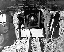 Photographie en noir et blanc. Dans ce qui semble être une entrée de mine, au milieu de la voie, deux hommes, dont l'un porte un costume militaire, relèvent un portrait dans son cadre.