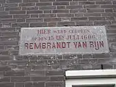 Photographie en couleurs. Plaque en béton dans un mur de briques rouges, sur laquelle on peut lire : "HIER werd geboren op den 15 den JULI 1606 REMBRANDT VAN RIJN"