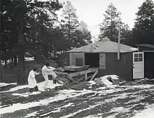 Une cabane entourée par des pins. Il y a de la neige sur le sol. Un homme et une femme en blouses blanches de laboratoire tirent sur une corde reliée à un petit chariot sur une plateforme en bois. Un large objet cylindrique est situé sur le chariot.