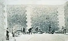 Photomontage en noir et blanc, nettement pâli, qui montre l'évacuation de cadavres étendus au sol, en arrière-plan, devant le haut mur de la cour intérieure d'une prison.