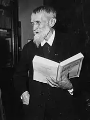 Photo en noir et blanc d'un vieillard vêtu de sombre, cheveux courts et barbe blanche, un grand livre ouvert à la main