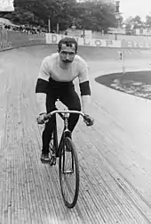 Photographie en noir et blanc d'un cycliste assis sur la selle de son vélo, en pull et pantalon, sur un vélodrome.