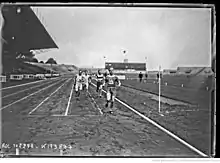 Photographie en noir et blanc d'un homme en tenue de sport, franchissant une ligne d'arrivée. Il s'agit de René Wiriath. Il porte le dossard no 90. En arrière-plan, un groupe de 3 coureurs se présente sur la ligne d'arrivée.