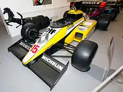 Photographie d'une monoplace de Formule 1 jaune, vue de trois-quarts, exposée dans un salon.