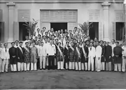 Un homme blanc en uniforme, entouré de plusieurs dizaines d'autres hommes, pour la plupart asiatiques, habillés à l'occidentale ou de manière traditionnelle, posant devant une maison