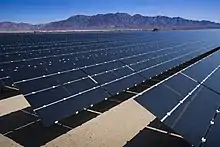 Plusieurs milliers de panneaux solaires installés dans un désert de sable. Au fond de l'image, une montagne rocheuse.