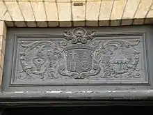 Une photo de l’imposte de la porte principale de l’église. Le blason de la basilique, encadré de ses attributs (pavillon et tintinnabule), est sculpté en bas-relief sur un panneau de bois sombre.