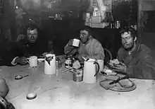 Photographie noir et blanc de trois hommes à table.