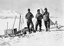 Photographie noir et blanc de trois hommes en tenue polaire, une tente derrière eux et un traineau à leur droite.