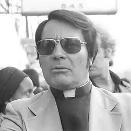 Photo en noir et blanc de Jim Jones, portant un vêtement de pasteur et des lunettes noires, devant une foule et un panneau qui indique Hotel.