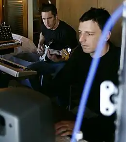 Photographie de deux hommes aux cheveux noirs et habillés en noir. L'un d'entre eux est en train de jouer de la guitare.