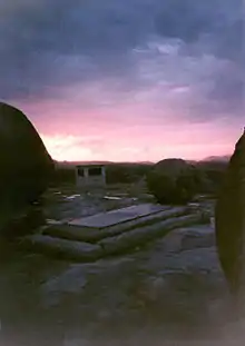 Photographie d'un lever de soleil sur un paysage montagneux avec des arbres sur l'horizon. Au premier plan se trouve une pierre tombale rectangulaire tandis qu'un monument massif est visible quelques dizaines de mètres derrière.