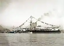 Une photo montrant un bateau à vapeur dans un port, décoré de drapeaux et une tourelle bien visible vers l'avant