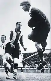 Photographie en noir et blanc d'un gardien de football, de dos, captant le ballon, devant des attaquants adverses.