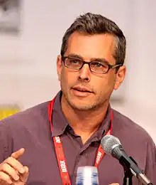 Photographie du buste d'un homme. Il porte un polo mauve, des lunettes et se trouve face à un microphone.