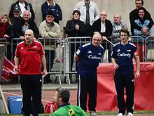 Trois hommes observent un match de rugby depuis le bord du terrain.