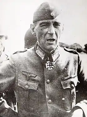 Reproduction d'une photo en noir et blanc montrant un homme, habillé en uniforme militaire d'officier allemand, avec une croix de fer. Son visage montre une expression fatiguée voire hébétée. Derrière lui, on devine un soldat américain avec son casque.