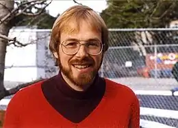 Portrait d'un homme blond, barbu, portant des lunettes, qui sourit en regardant l'objectif.