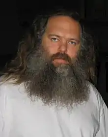 Homme avec une longue barbe grise portant un t-shirt blanc.