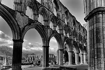 Photographie noir et blanc d'uné église en ruine à travers les arcades de laquelle on peut voir d'autres ruines en arrière-plan.