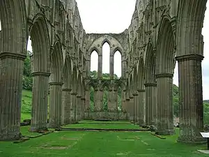 Photographie d’une nef d'église dont il ne reste que les arches latérales, sans bas-côtés, ni voûtes, ni dallage, ce dernier ayant été remplacé par de l'herbe.
