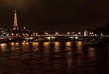 Photo prise de nuit avec pont et Tour Eiffel éclairés au milieu, entre un ciel noir et des eaux sombres reflétant les lumières.