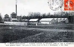 Carte postale en noir et blanc représentant un ensemble de bâtiments dominés par une cheminée d'usine en briques.