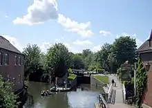 Canal s'écoulant depuis le photographe et qui se divise en deux canaux avec celui de droite arrivant aux portes de l'écluse. A gauche, un bâtiment et à droite un sentier et un pub.