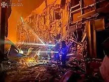 Photographie en couleurs. Des pompiers inspectent, lampe à la main, un bâtiment en ruine. Arrière-plan couvert par un écran de fumée et l’obscurité. Le logo en haut à gauche indique que la photographie provient d’une source officielle.